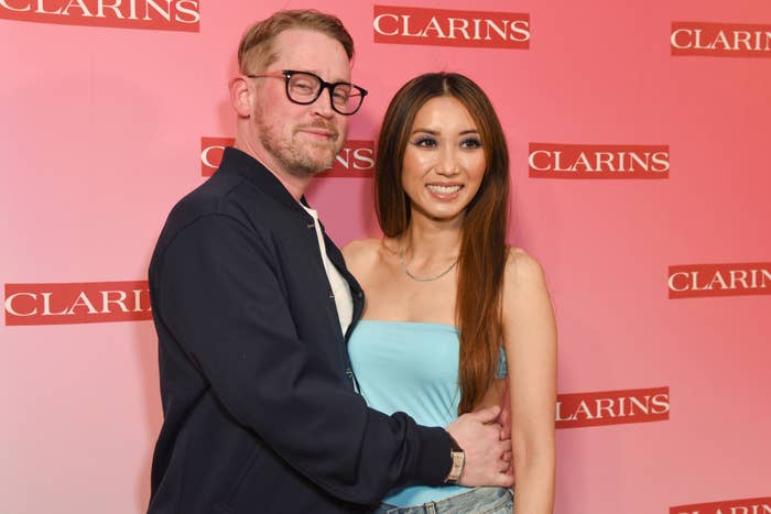 Two people posing together, one in a casual jacket and glasses, the other in a strapless dress, at a Clarins event