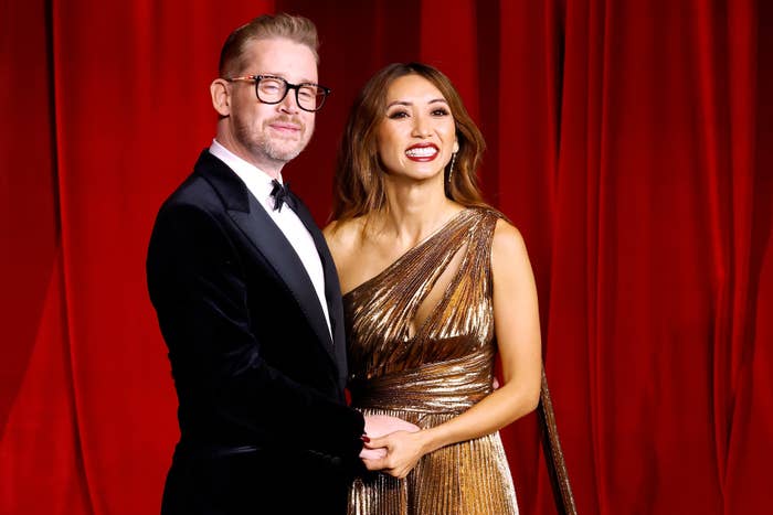 A couple poses on the red carpet; the man wears a velvet tuxedo, and the woman is in a shimmering metallic gown