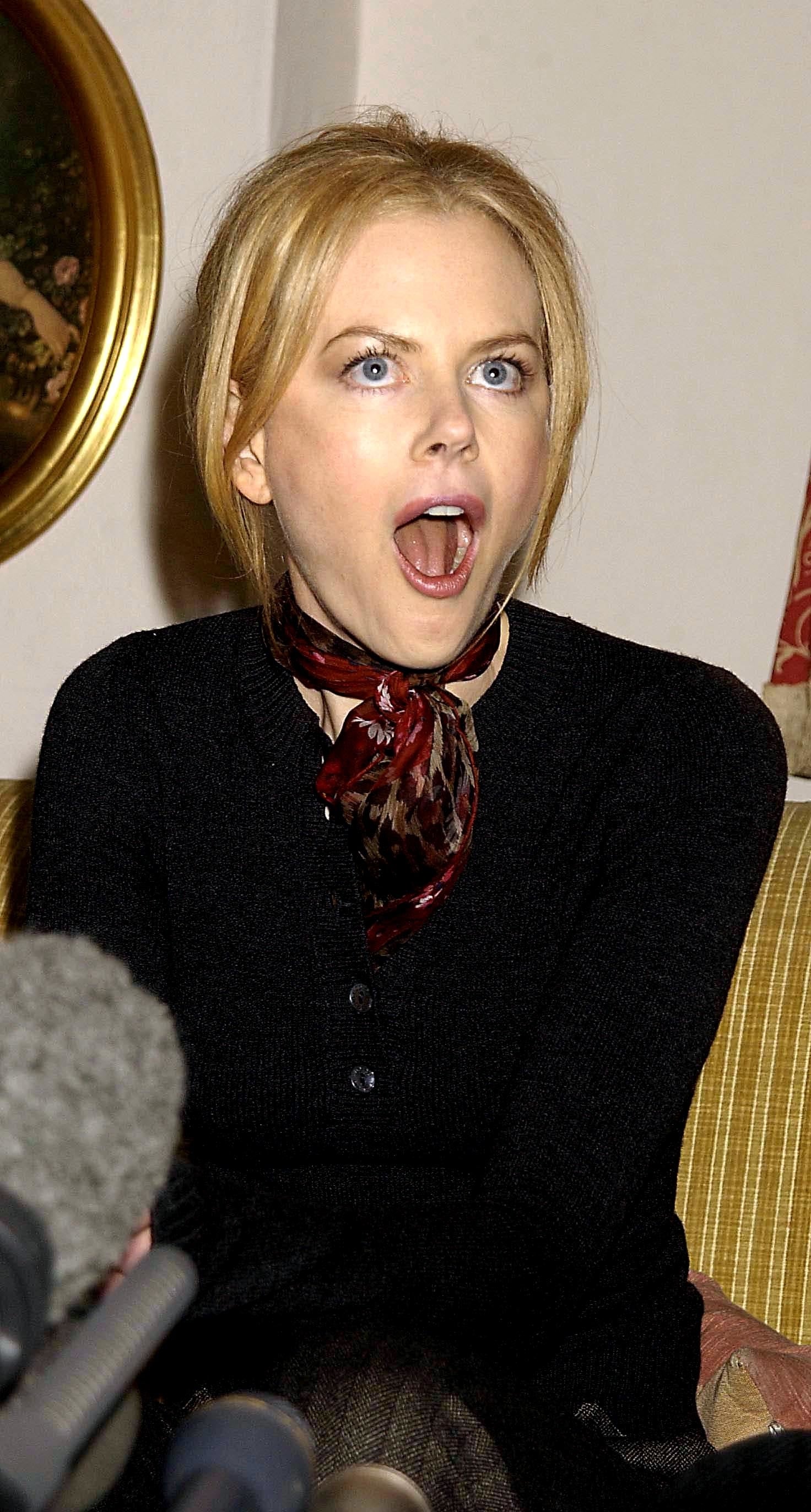 Nicole Kidman sitting on a couch, looking surprised with mouth open, wearing a dark top and a patterned scarf around their neck. Microphone visible in foreground
