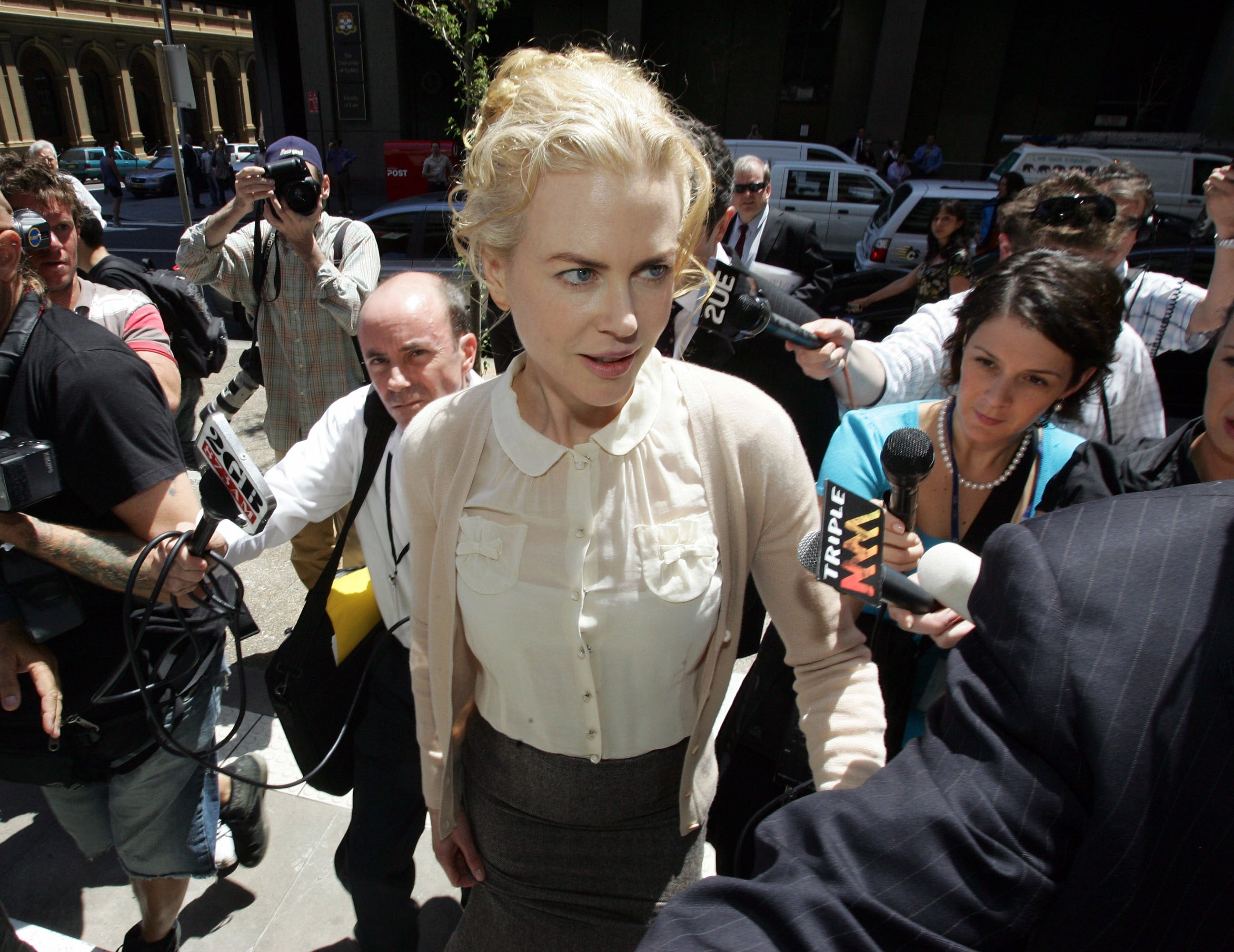 Nicole Kidman surrounded by media, wearing a blouse and skirt, walks through a crowded area with microphones and cameras around