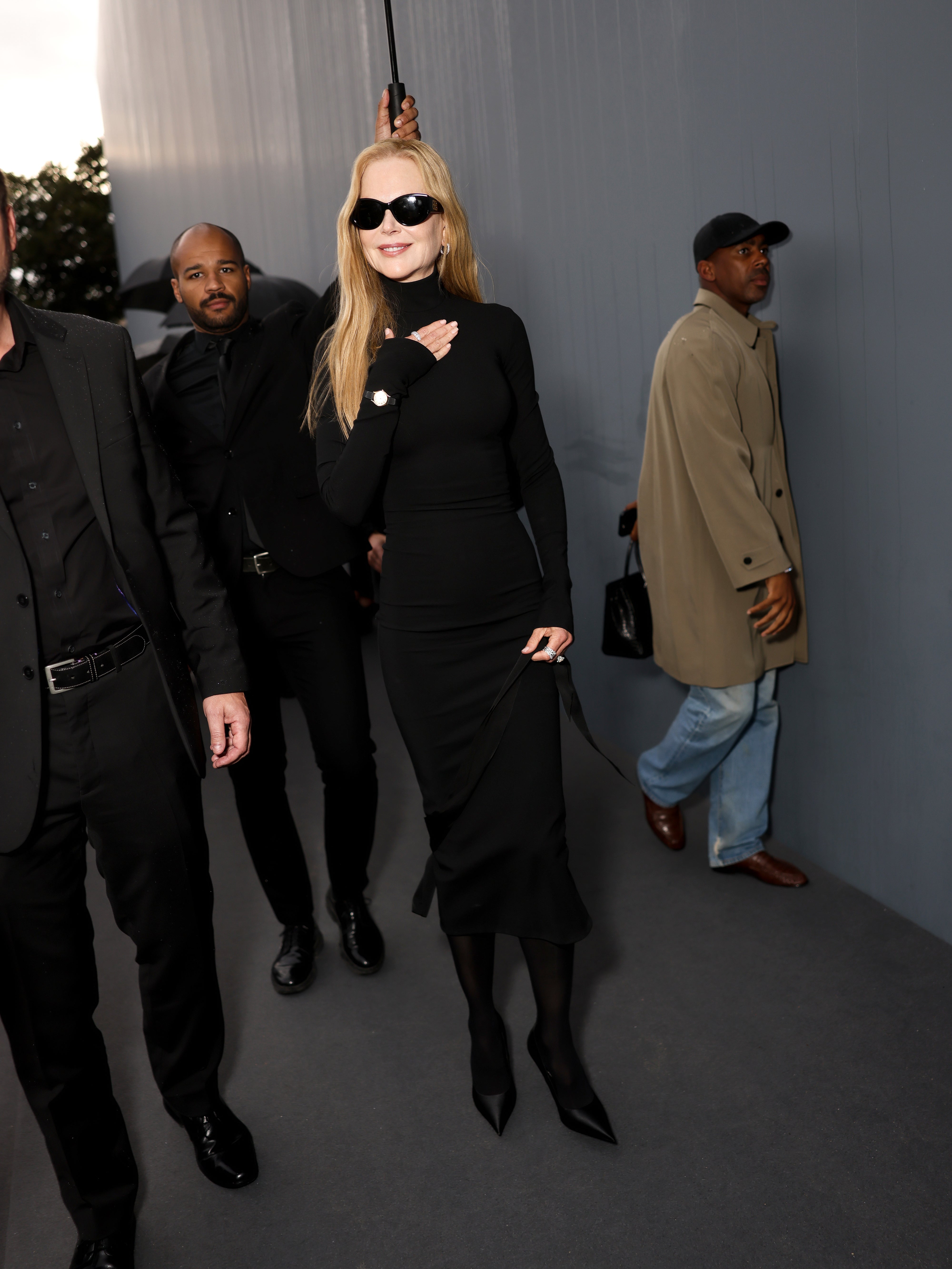 Nicole Kidman in a stylish black dress, sunglasses, and heels walking with security. Another person in a trench coat is nearby