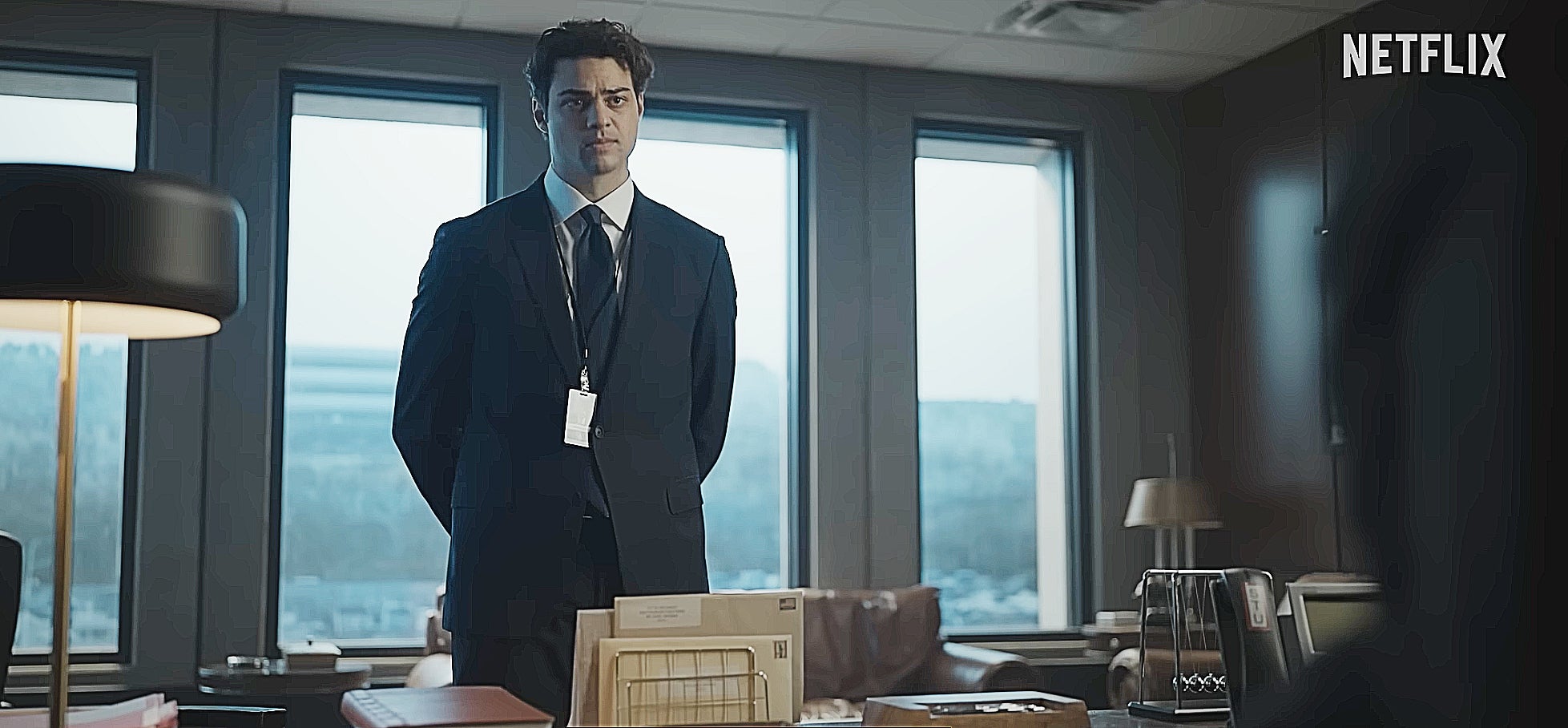 Man in a suit stands behind a desk in an office setting, Netflix logo in corner