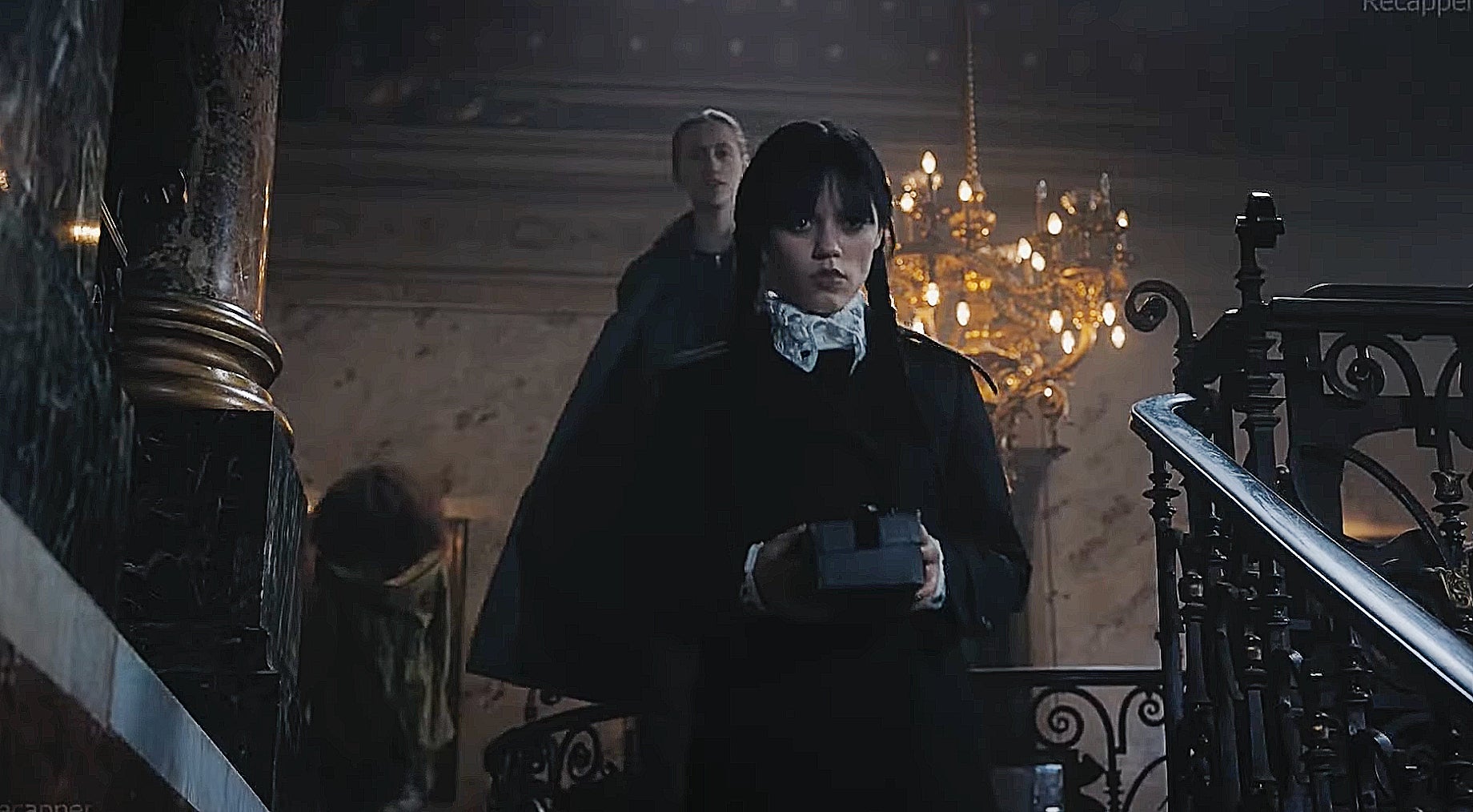 Person in gothic attire with a white ruffled collar holds a box, standing on a grand staircase with a chandelier overhead