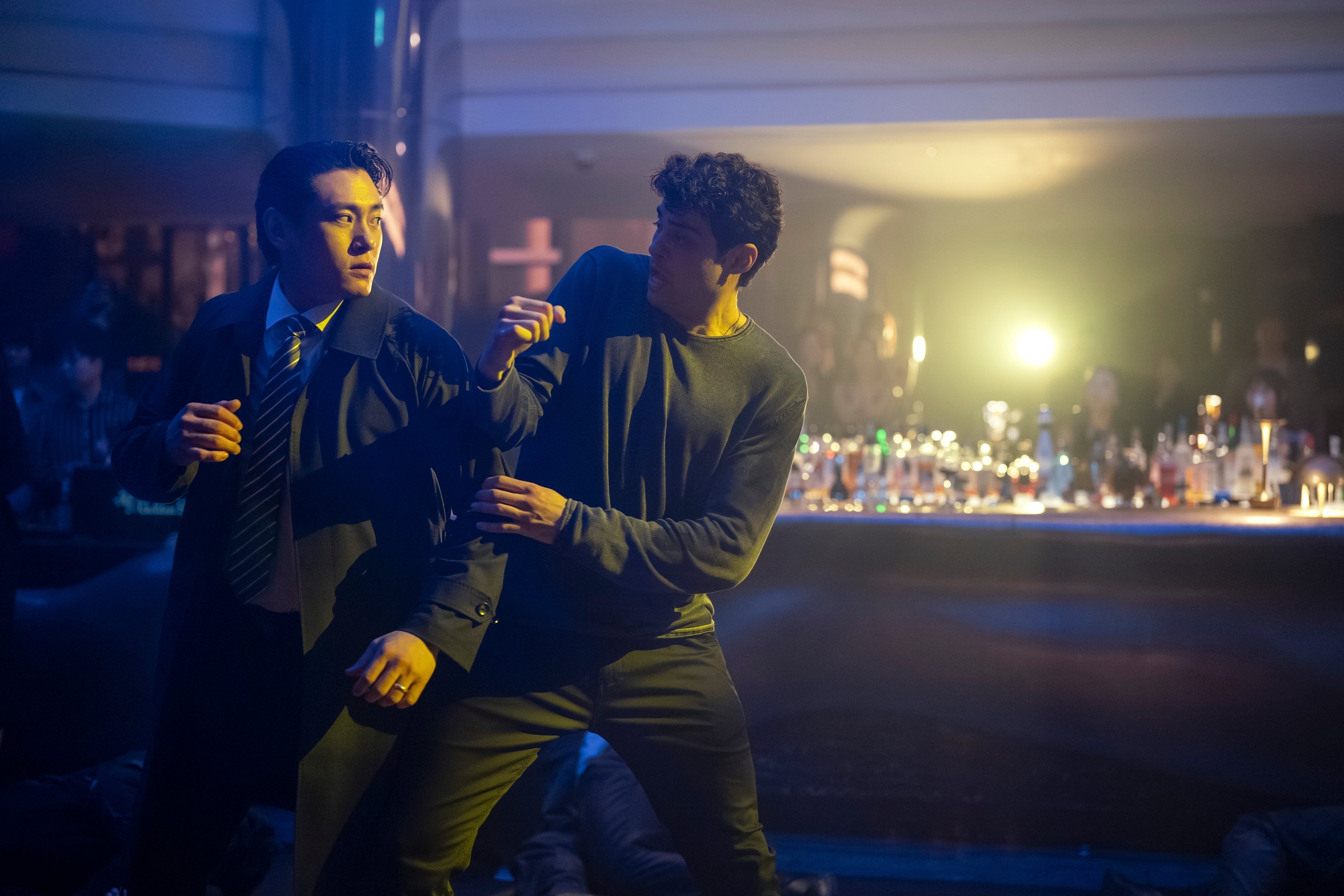 Two people in a tense confrontation in a dimly lit bar setting. One wears a suit, the other a casual long-sleeve shirt