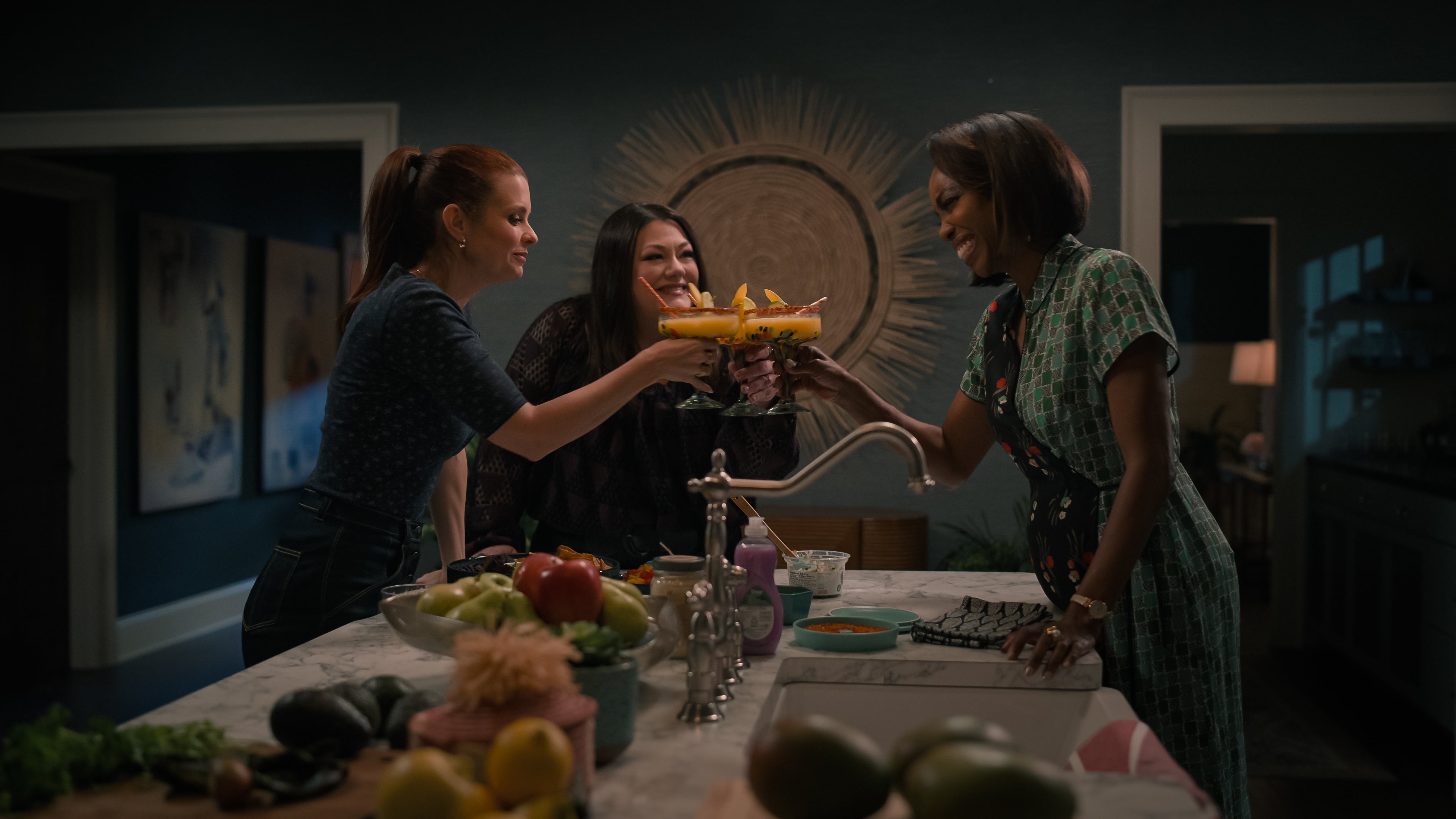 Three women in a kitchen clink cocktail glasses while smiling, surrounded by fruits and vegetables on the counter