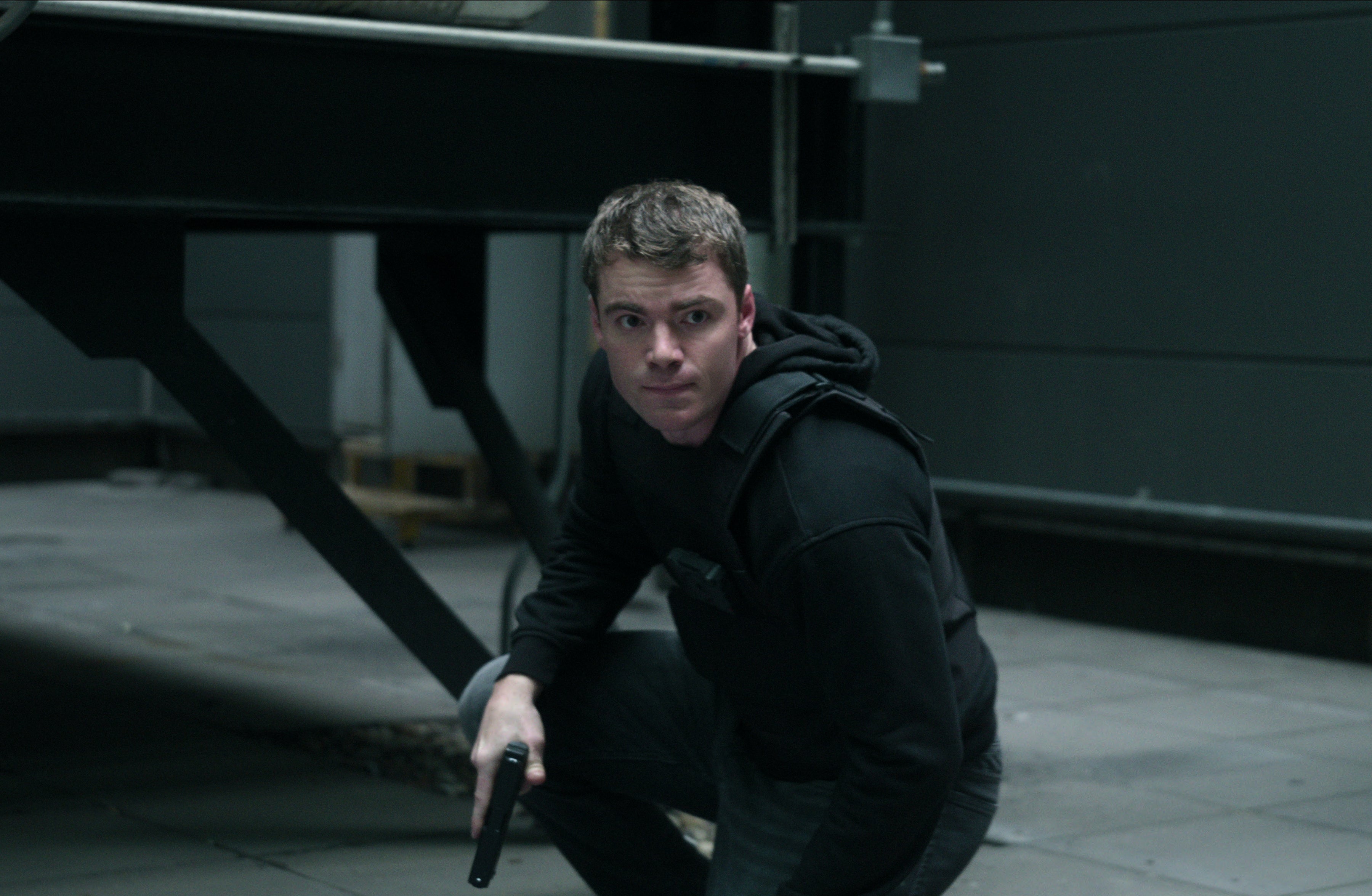 Peter Sutherland from "The Night Agent" kneeling with a focused expression, holding a weapon