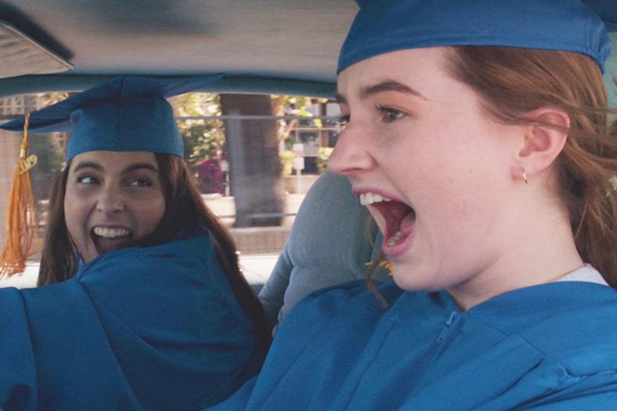 Beanie Feldstein and Kaitlyn Dever wearing graduation caps and gowns joyfully celebrate in a car