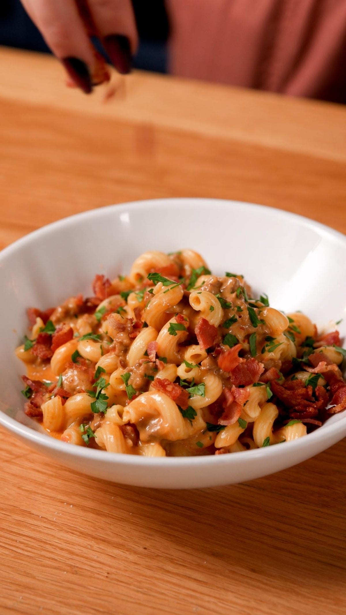 A bowl of pasta with creamy sauce, topped with crispy bacon bits and fresh parsley, is being lightly garnished by a hand