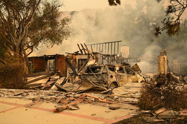 House and car destroyed by a wildfire; debris and smoke fill the scene, with trees in the background