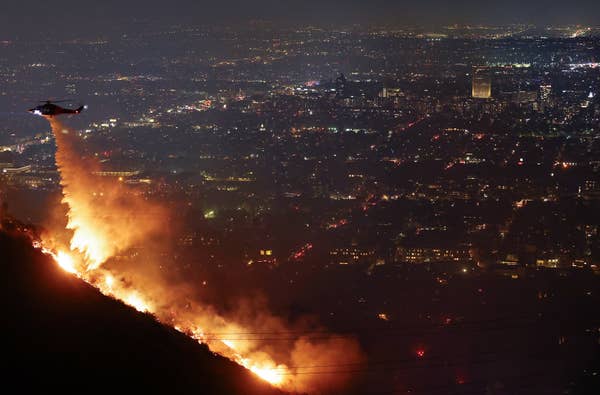 A helicopter drops water on a hillside wildfire at night, with a cityscape visible in the distance