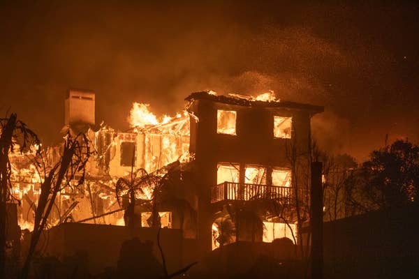 A large house engulfed in flames at night, with bright fire illuminating the building and surrounding area