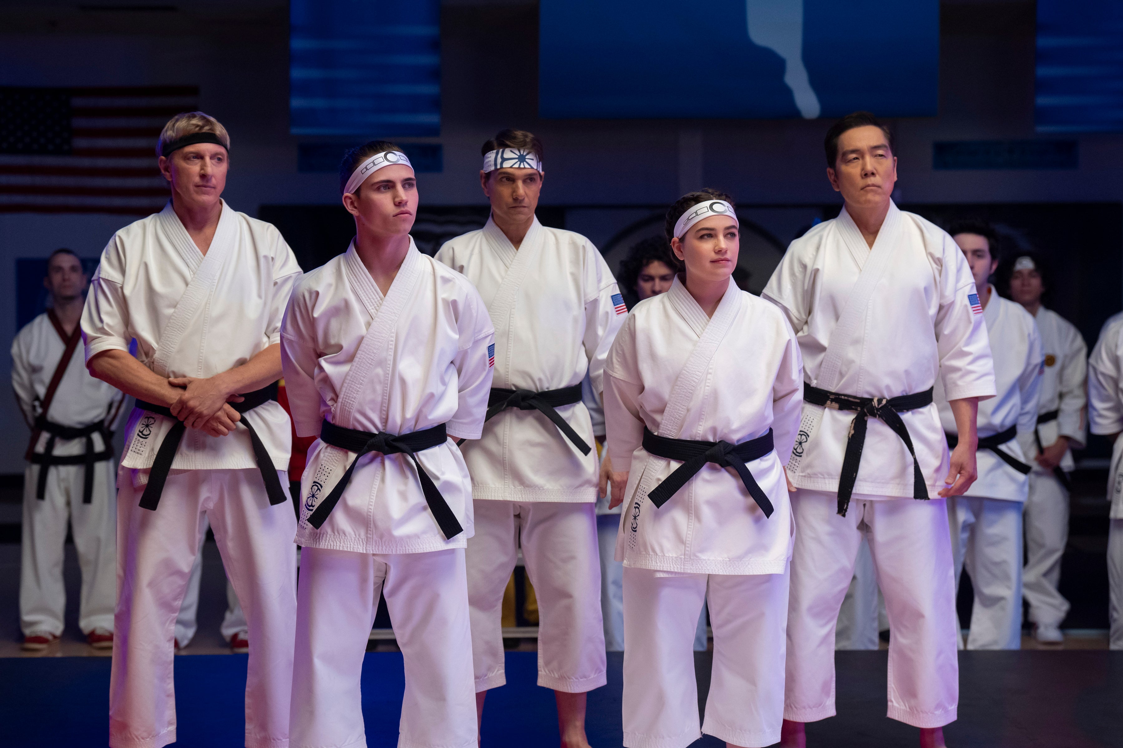 A group of five people in karate uniforms with black belts stand in a martial arts setting, appearing focused and attentive