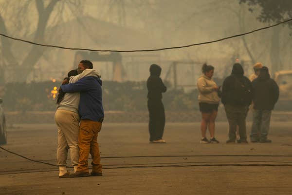 People hugging on a smoky street, others standing in the background, possibly after a disaster or emergency