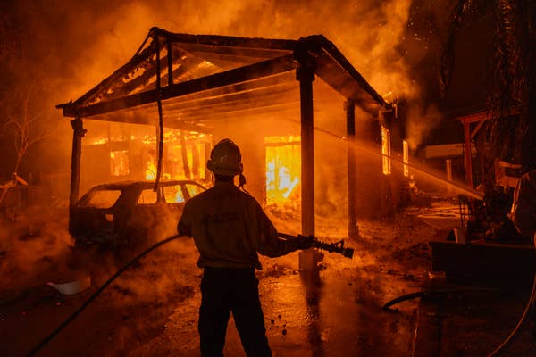 Firefighter battling a large house fire at night, spraying water onto the flames engulfing the structure and a nearby car