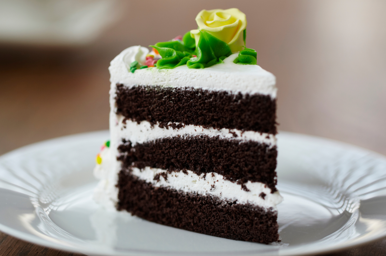 Slice of chocolate cake with vanilla frosting and flower decorations on top, placed on a plate
