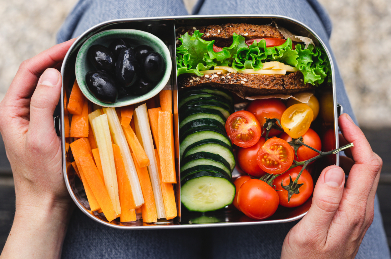 Person holding a lunchbox with a sandwich, sliced cucumber, cherry tomatoes, carrot sticks, and a small bowl of black olives