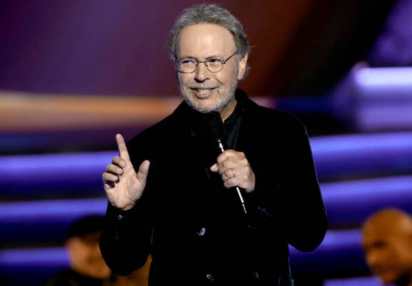 Person in a dark velvet suit holds a microphone, gesturing while speaking in front of an audience