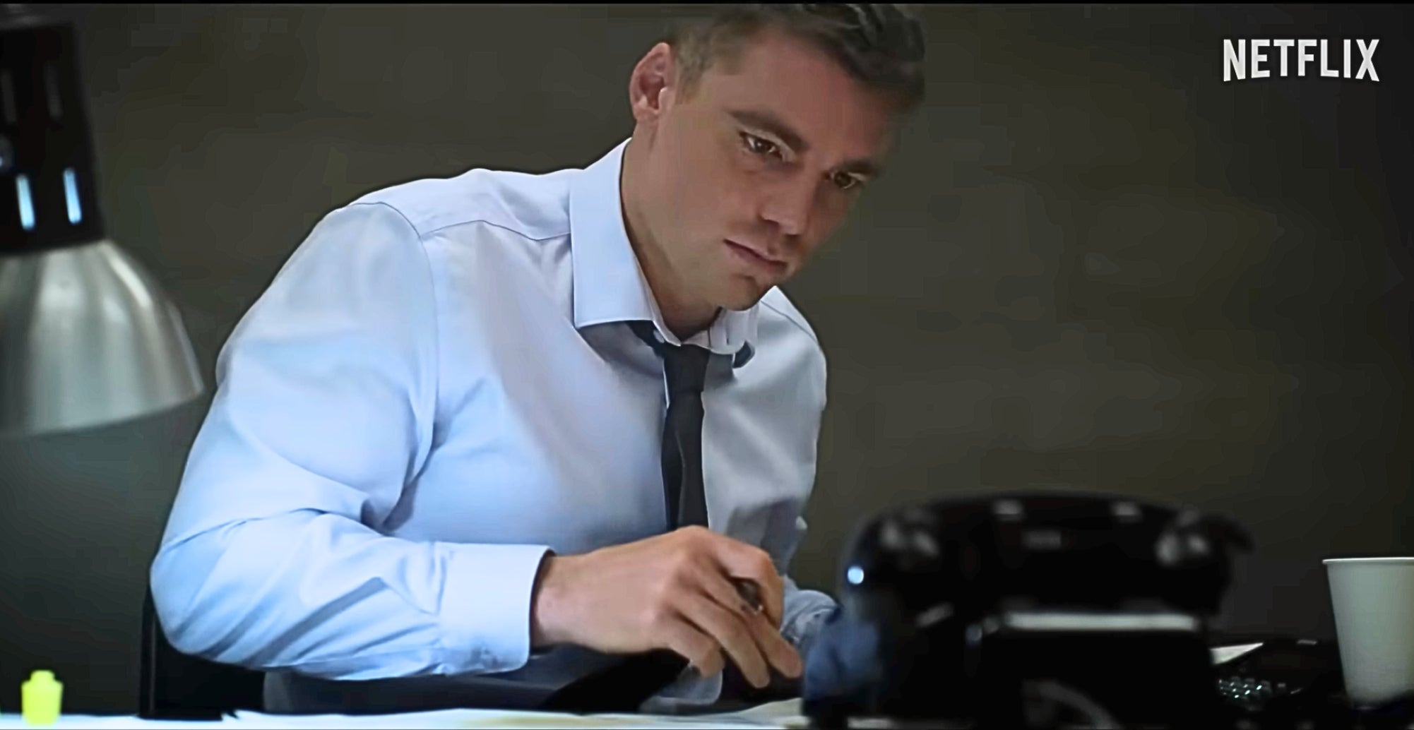 Person in office setting, wearing a white shirt and tie, looking intently at papers on a desk