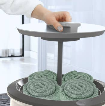 Hand lifting lid of a compact towel warmer with neatly rolled towels inside, suitable for bath buying articles