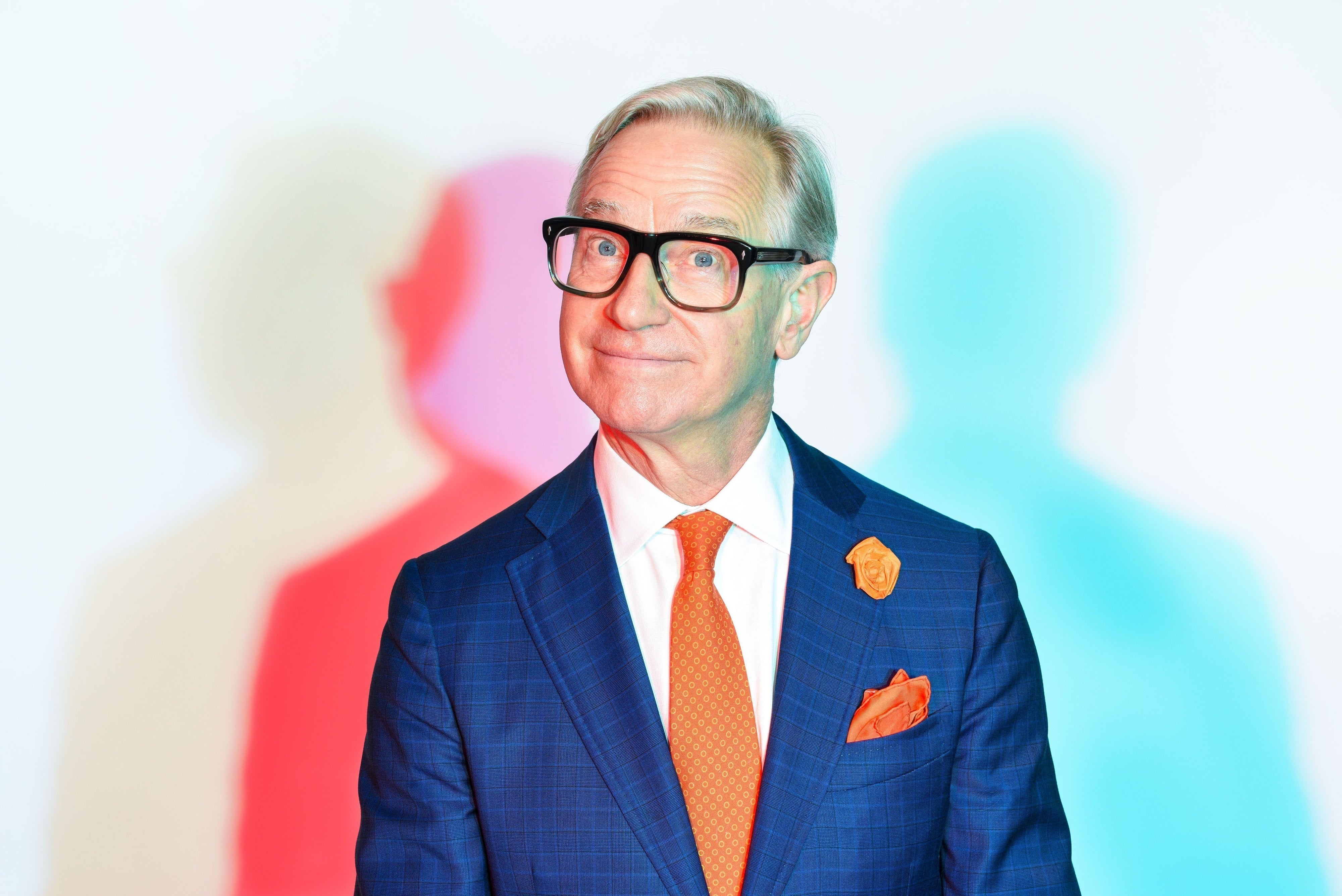 Paul Feig in a suit with glasses poses in front of colorful shadows, wearing a patterned tie and pocket square