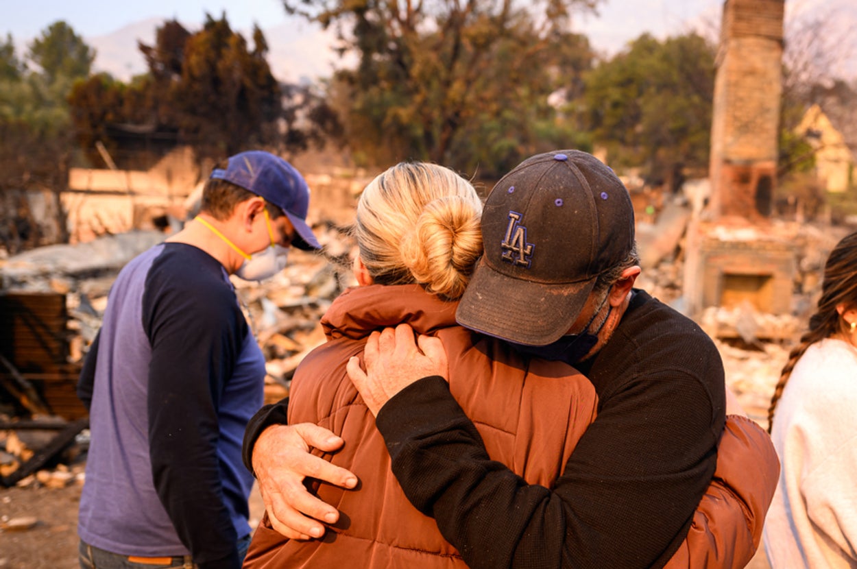 People embrace in front of a fire-damaged home, with rubble around them. Others look on, appearing concerned