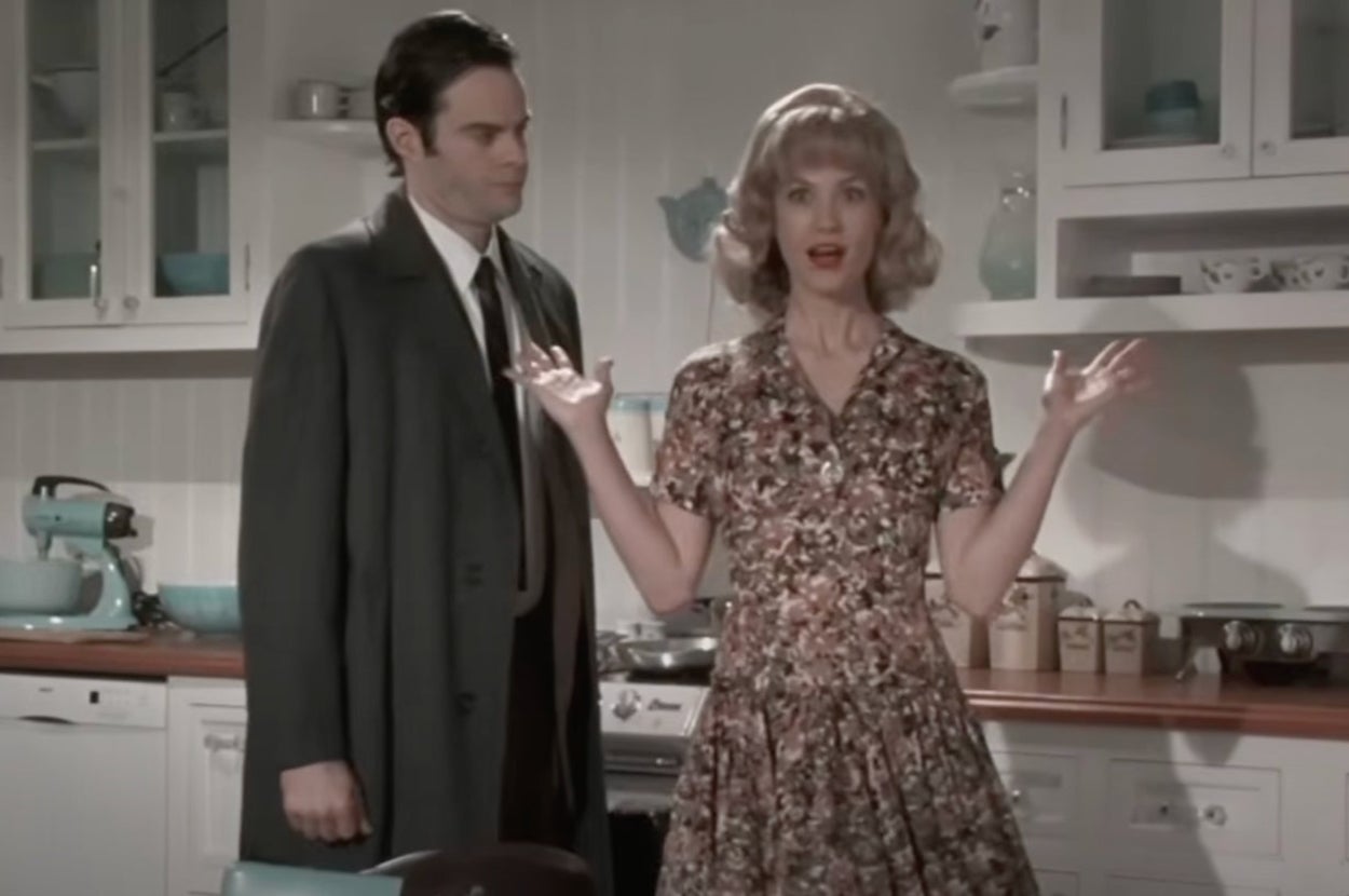 Two actors in a vintage kitchen set; the woman wears a floral dress, gesturing with surprise, while the man in a suit watches her