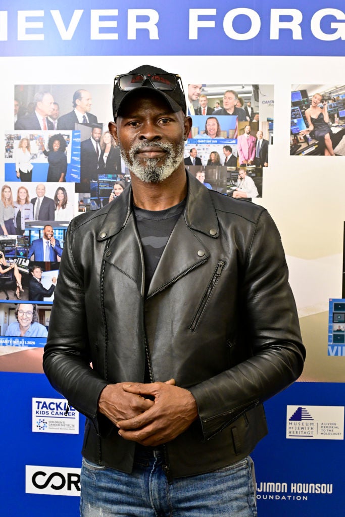 Person wearing a leather jacket and jeans at a celebrity event. Various photos and logos are displayed in the background