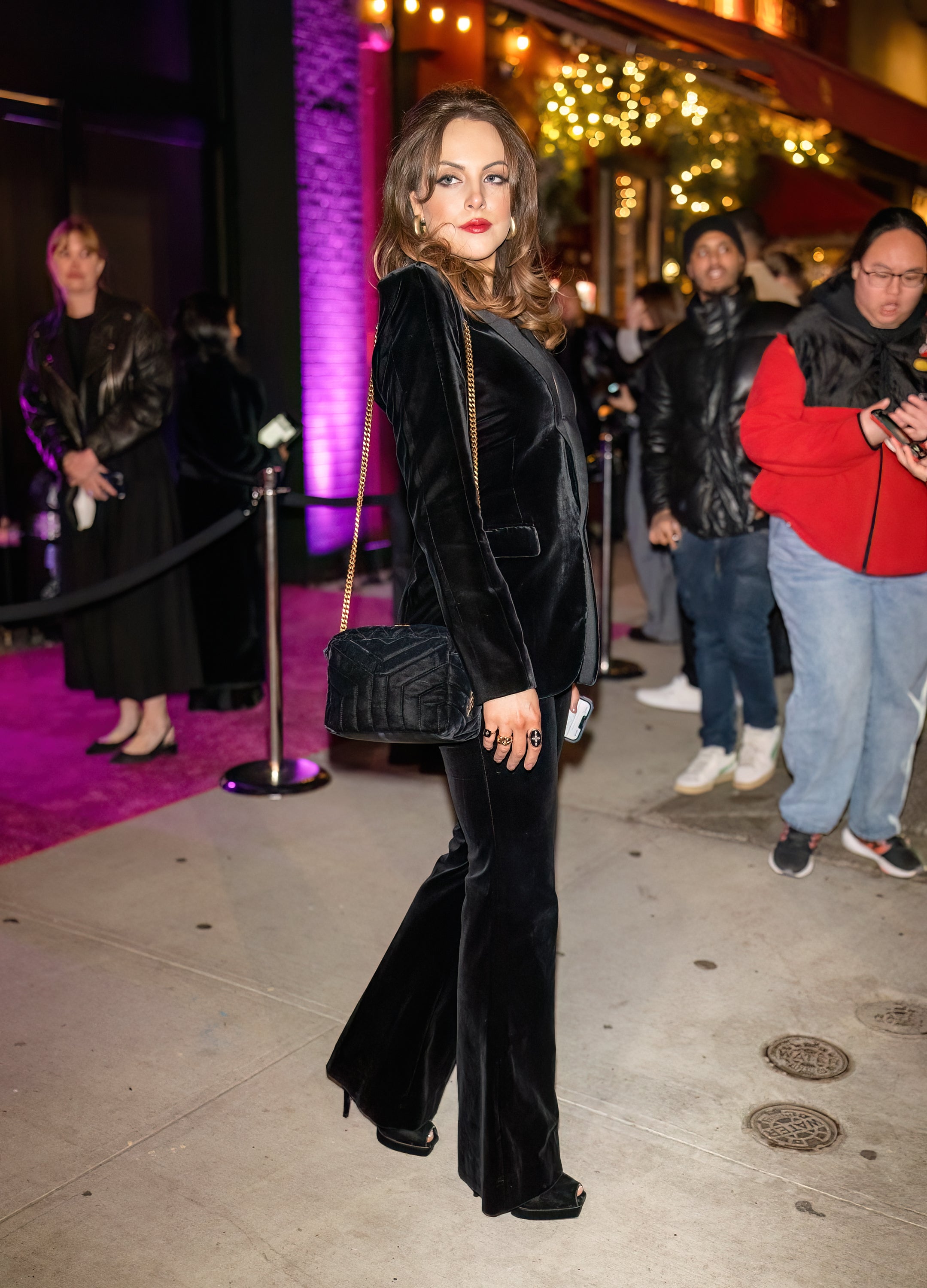 Elizabeth Gillies in a stylish velvet pantsuit poses confidently on a city sidewalk at a nighttime event. Crowd and festive lights in the background