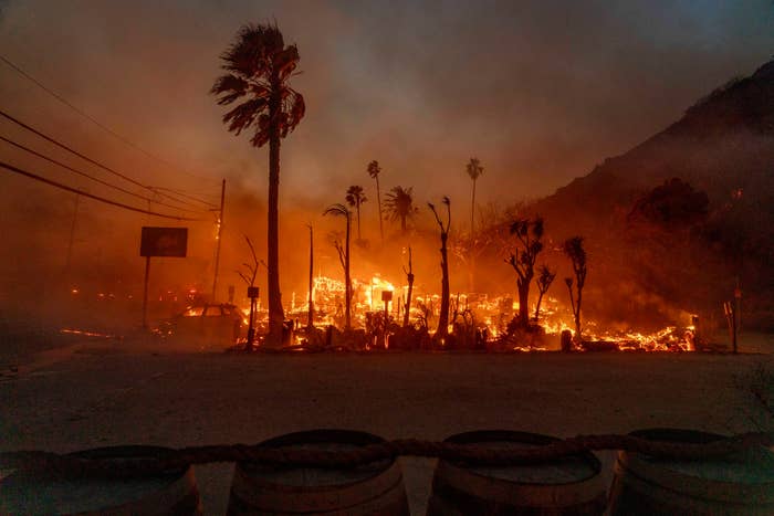Wildfire rages through a landscape, with intense flames engulfing trees and structures, creating a dramatic and destructive scene