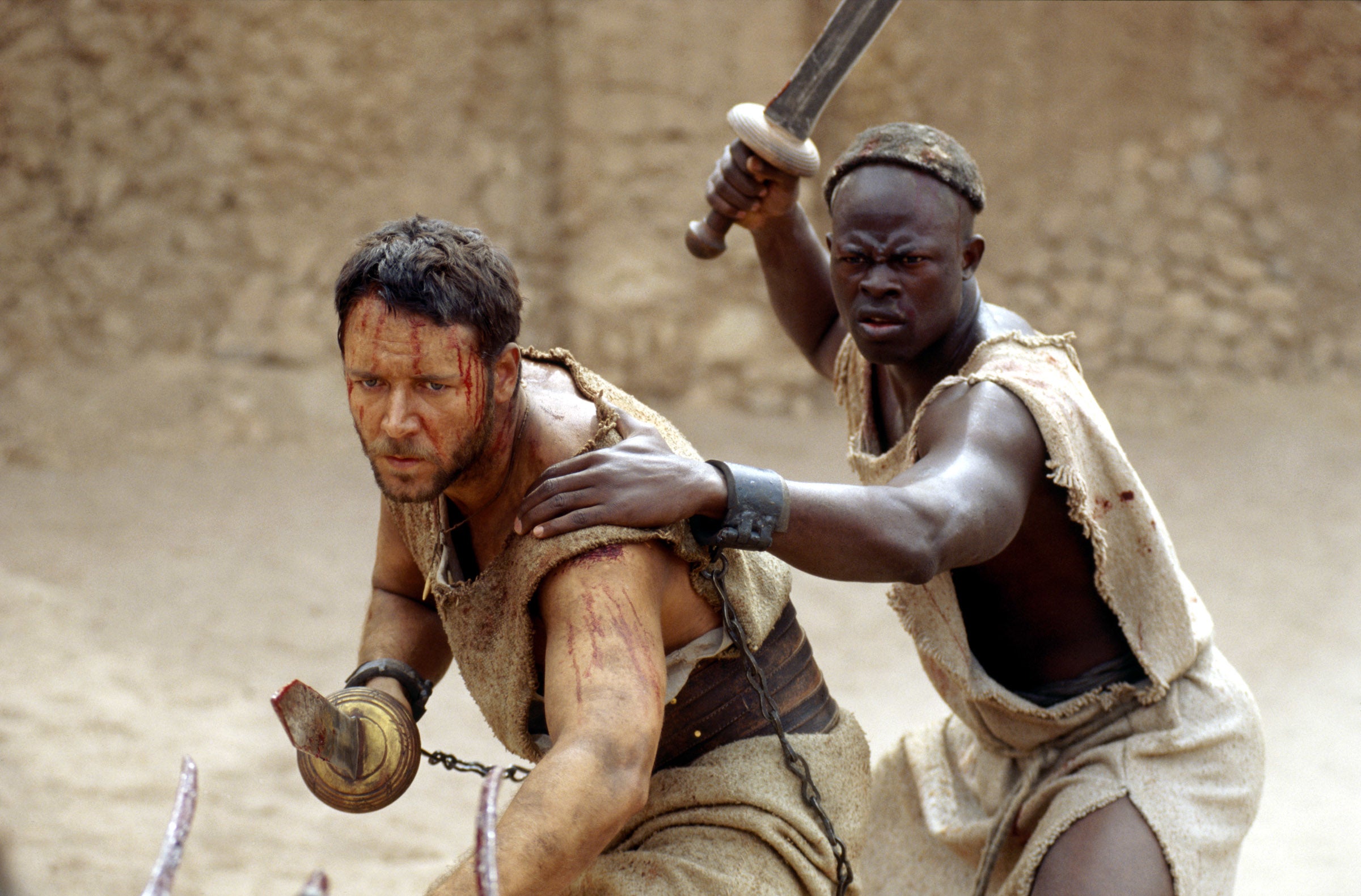 Two actors in a desert scene dressed as gladiators. One holds a sword and shields the other. They look intense and battle-worn