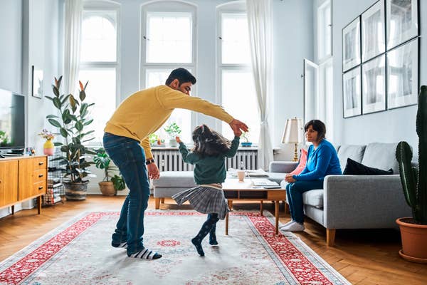 A family dances in a cozy living room; father and daughter twirl, mother watches while seated on sofa