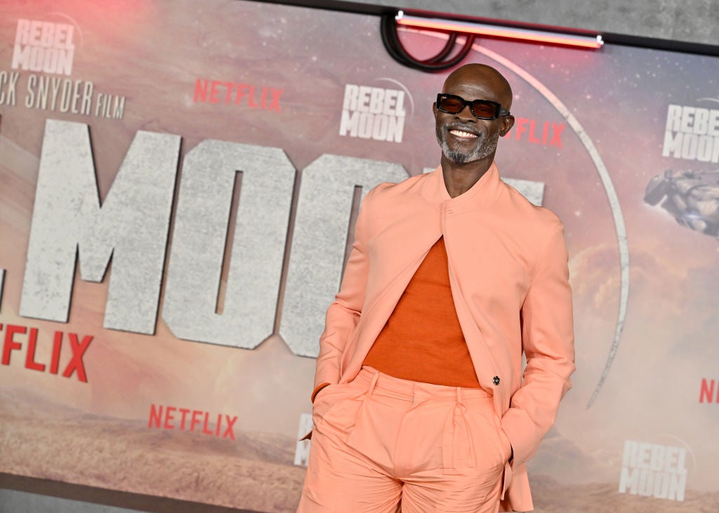 Person in stylish tan suit and sunglasses at a Netflix event with large "Rebel Moon" backdrop