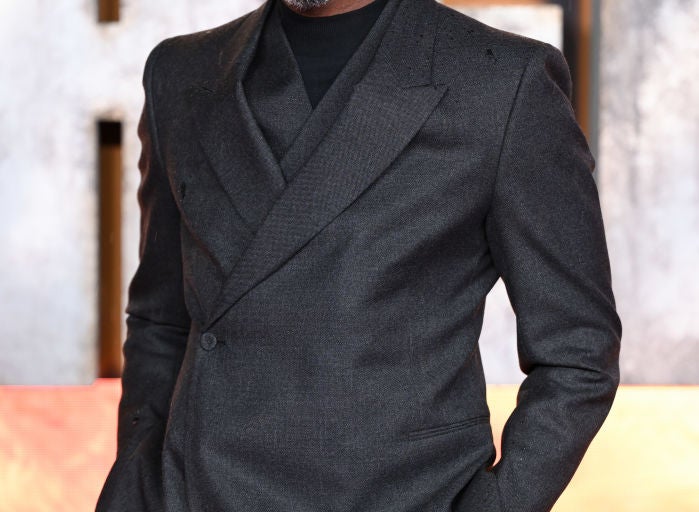 Man in a sharp, dark suit on a red carpet, smiling confidently with hands in pockets