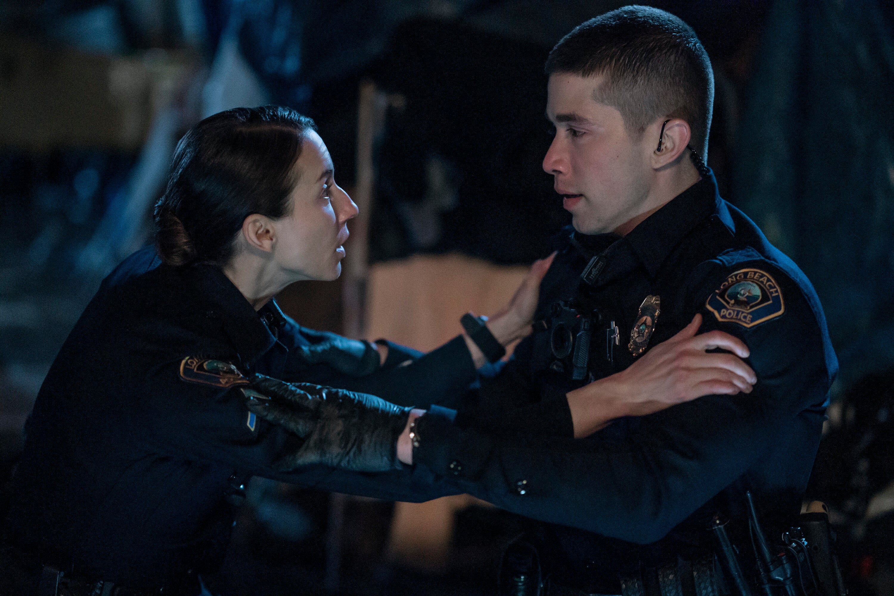 Two actors in police uniforms appear in an intense scene, one holding the other's shoulders, conveying urgency or concern