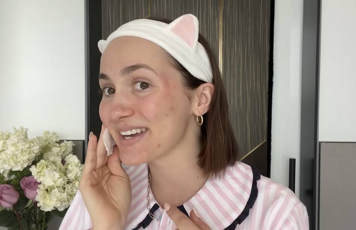 Celebrity uses a cotton pad on her face, wearing a headband with cat ears and a striped pajama top. Cream flowers are in the background