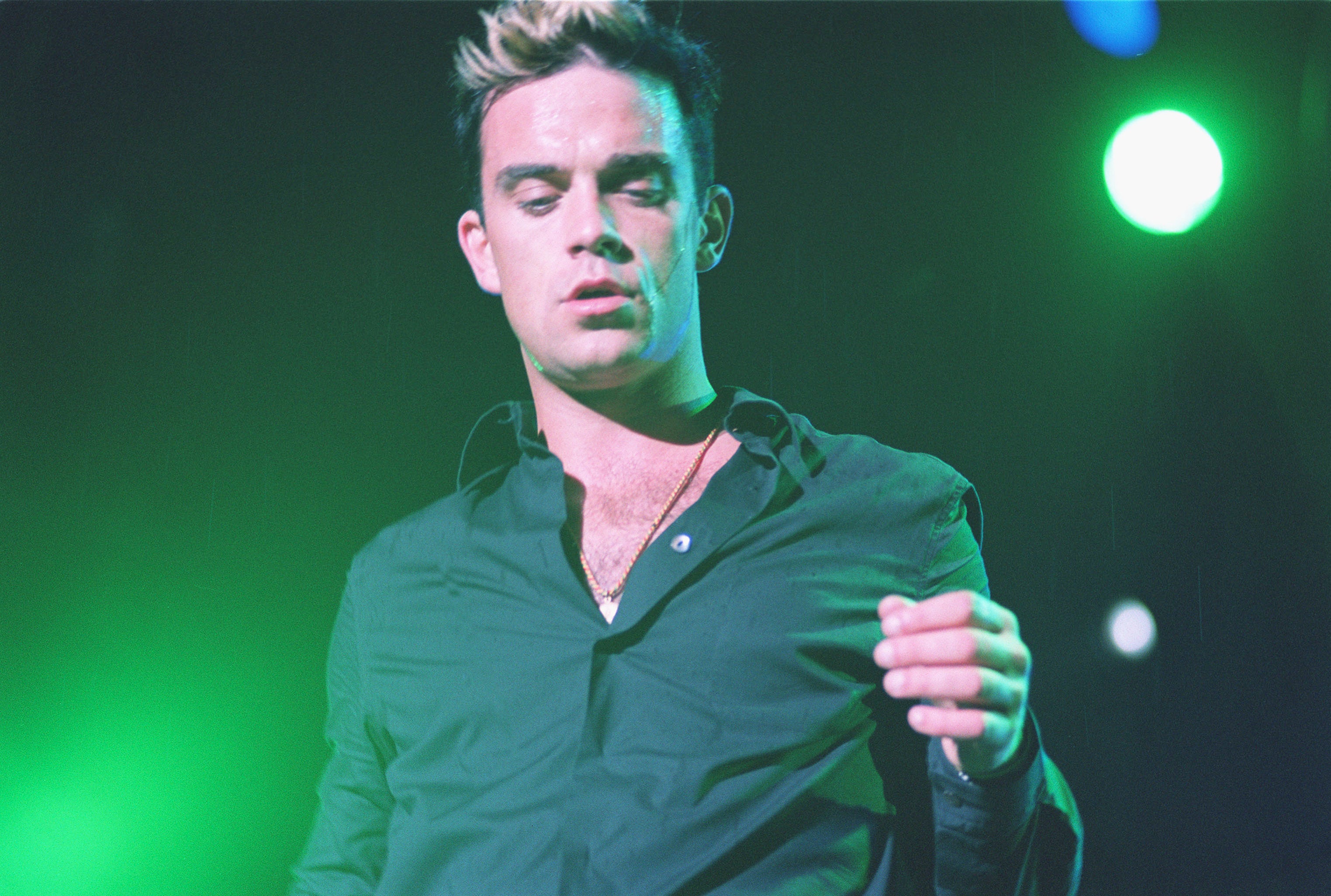 Robbie on stage, wearing a dark shirt, performing with lights in the background
