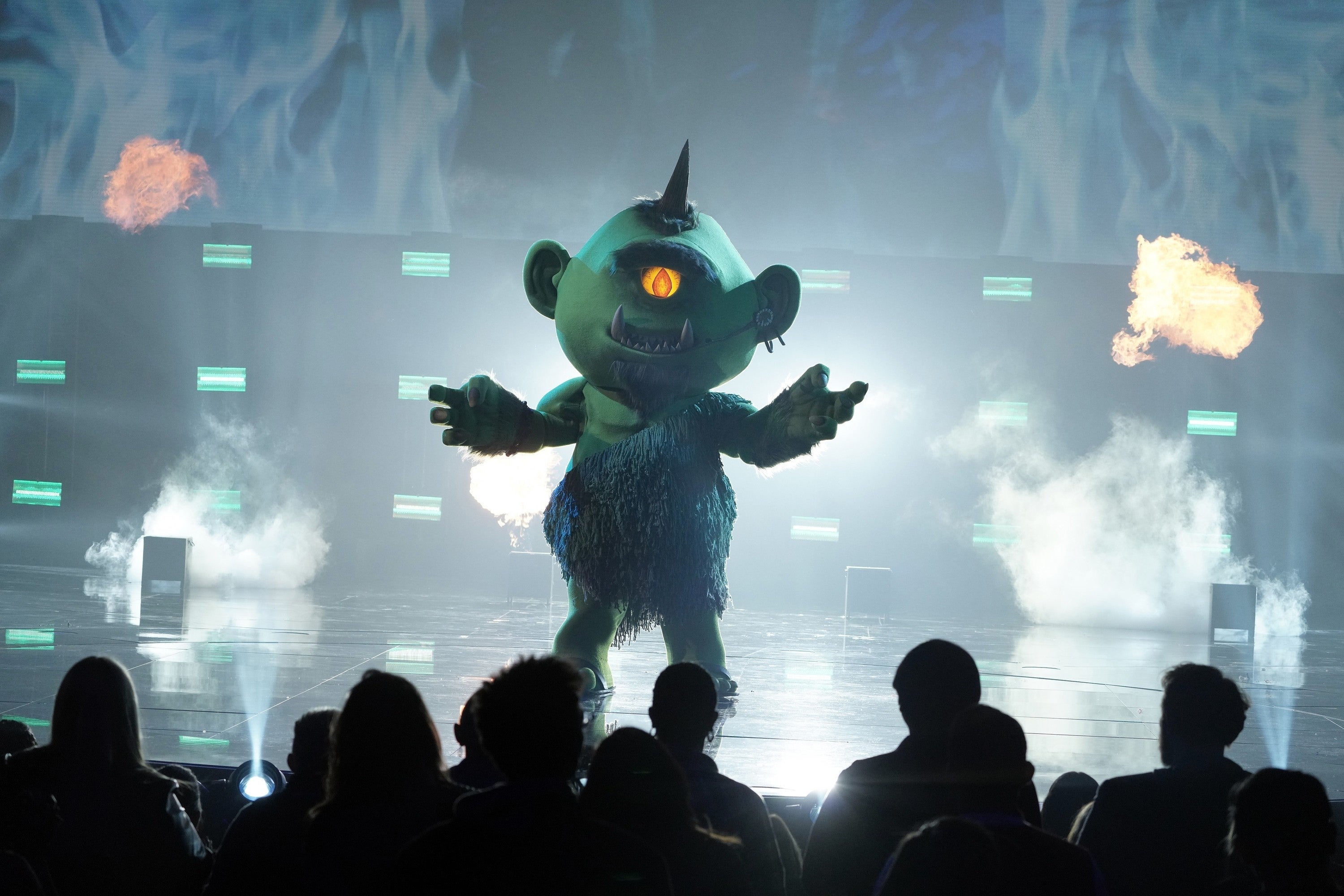 Costumed performer in a green, furry monster suit with one eye dances on stage, backlit by dramatic lights and surrounded by mist and flames. Audience watches