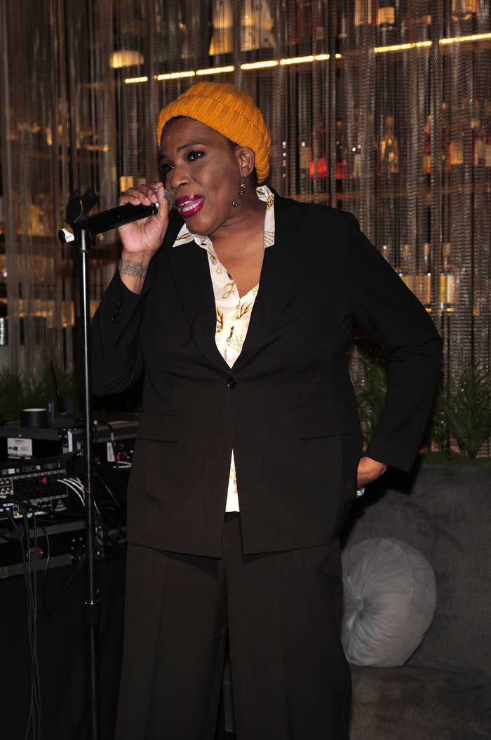 Macy Gray singing on stage, wearing a yellow beanie, a black suit with a white shirt, holding a microphone, in a warmly lit venue