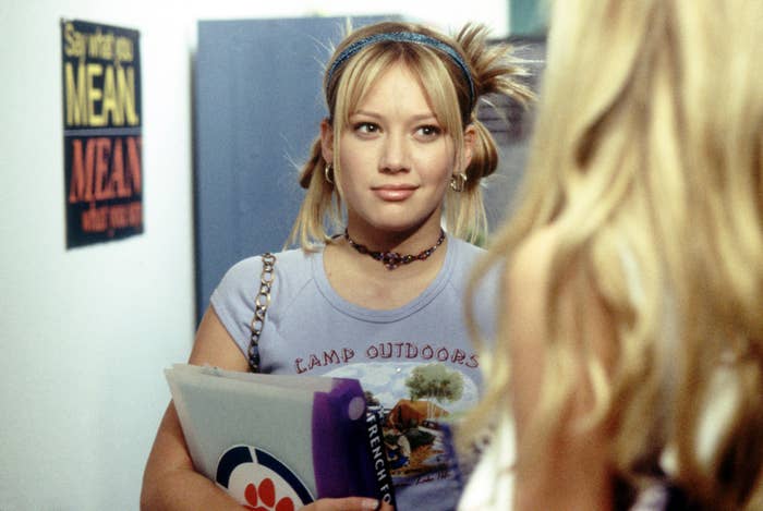 Person in a casual outfit with a graphic T-shirt and headband, holding a folder, looks at another person. A poster says, "Say what you mean."