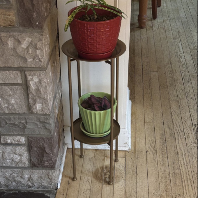 Two-tiered plant stand with potted plants: a red pot on top with a leafy plant and a green pot below, standing on a wooden floor by a stone wall