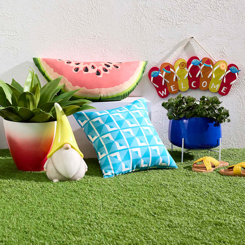 A garden scene with a watermelon pillow, a geometric blue cushion, potted plants, a gnome, a welcome sign, and yellow sandals on grass