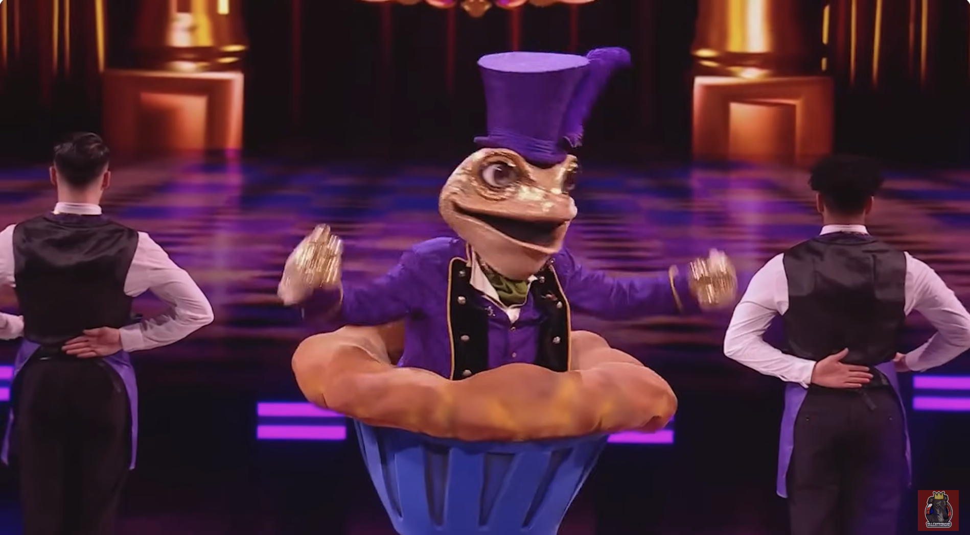 Person in elaborate frog costume with sparkly top hat performs on stage with two backup dancers in black vests