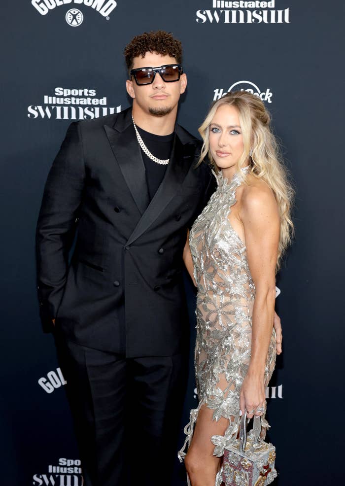 Two people pose on a Sports Illustrated Swimsuit event red carpet. One wears a black suit and sunglasses, the other a shiny, floral-patterned dress