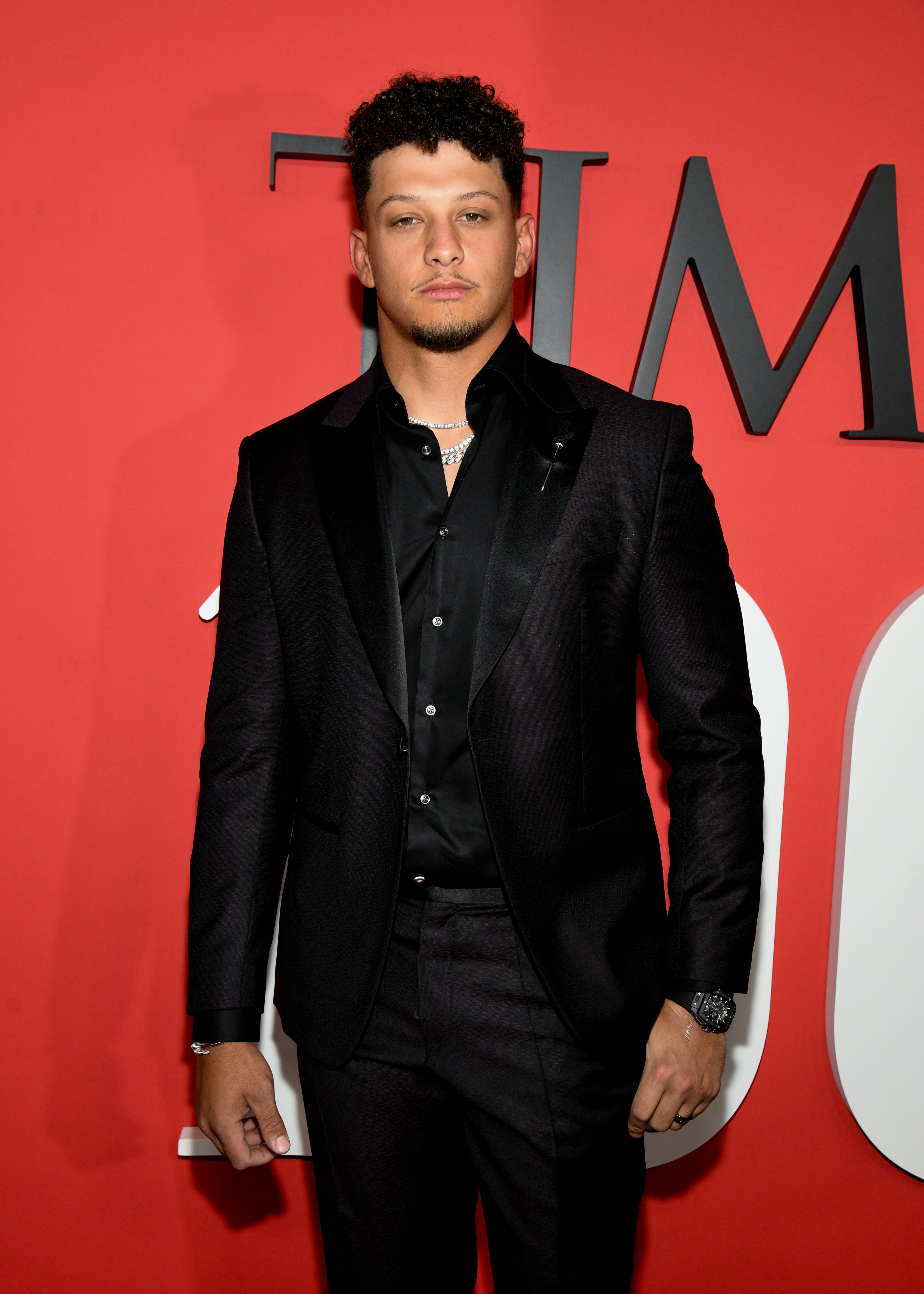 Person in a sleek black suit, standing in front of a red backdrop at a formal event