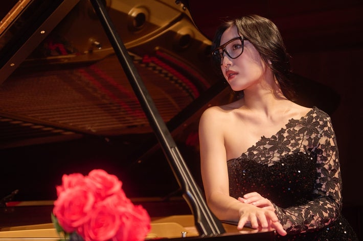 Woman in an elegant, one-shoulder lace evening gown stands beside a grand piano with roses on it