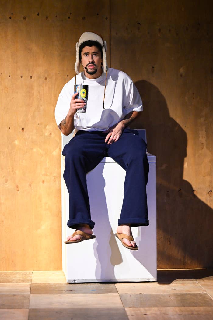 Bad Bunny sitting on a washing machine, wearing a casual white t-shirt, navy pants, flip-flops, and a warm hat, holding a beverage can