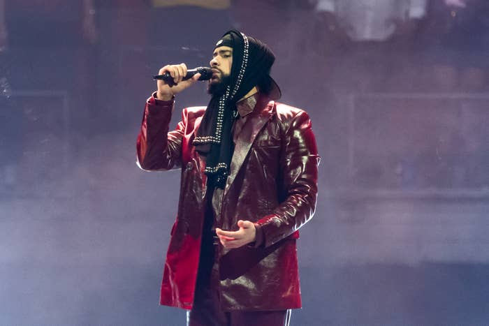 Bad Bunny in a shiny, leather-like jacket and hooded headpiece sings into a microphone on stage