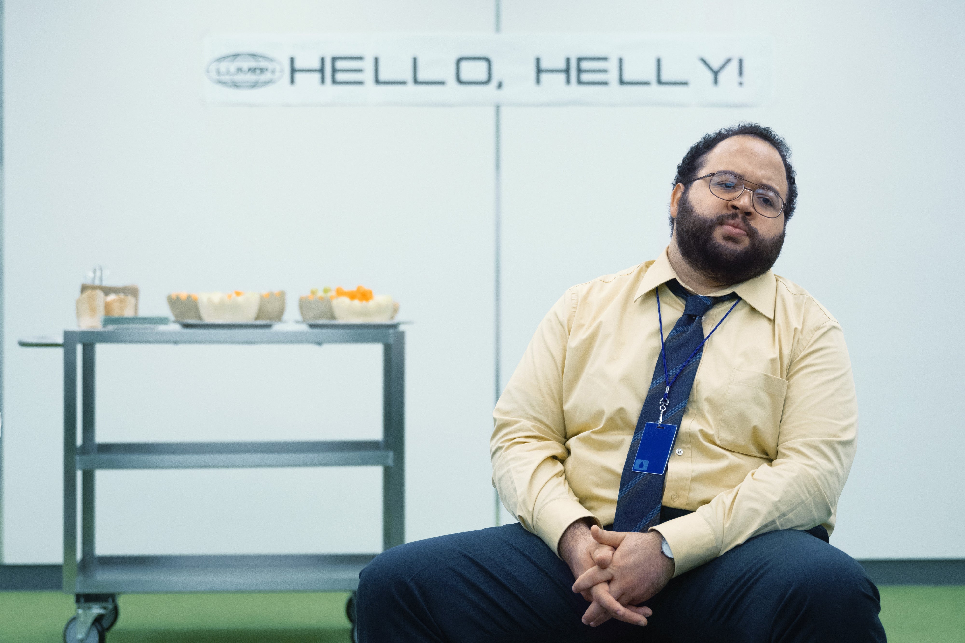Person in office attire sits in front of a cart with snacks, next to a "Hello, Helly!" sign. The scene suggests a workplace setting