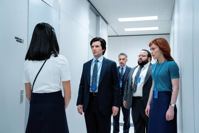 Five people, dressed in business attire, stand in a white hallway, facing one woman whose back is turned. Scene suggests a serious discussion