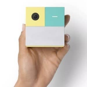Hand holding a small, quadrate camera with a minimalist design, featuring a lens and a fastener connected the apical section